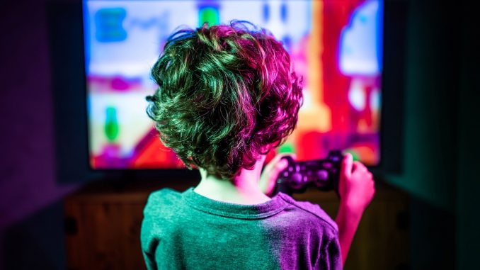 developpement-enfant-jeux-videos Un jeune garçon, manette à la main, jouant à un jeu vidéo ultracoloré sur un large moniteur TV.
