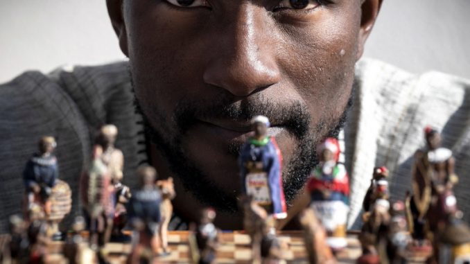 Teddy Kossoko, assis devant un jeu d’échecs avec des pions à l’effigie de personnages africains.