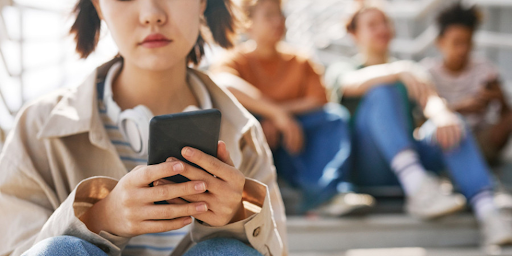 temps passé sur smartphone Une adolescente en train d'utiliser son smartphone et derrière des jeunes garçons qui discutent